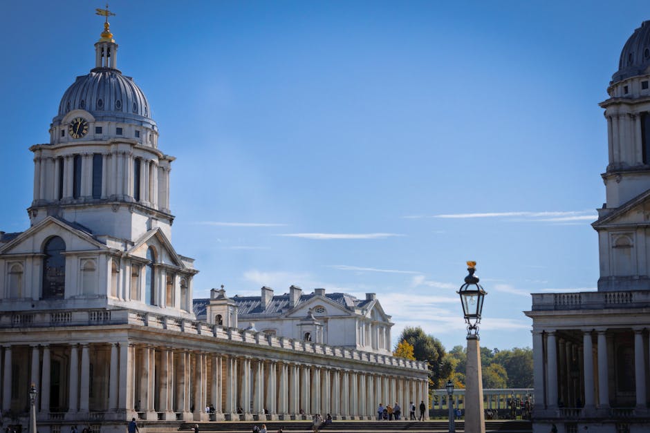 A historic domed building with classical architectural features, including columns and a decorative balustrade, situated on a grassy area with a fence around it. To the left, modern high-rise office b