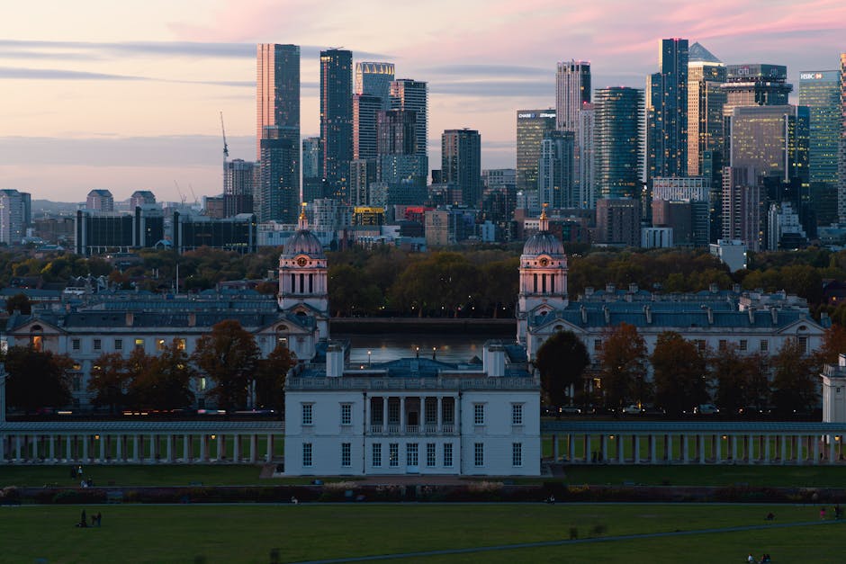 A large white historic mansion with symmetrical architecture, featuring multiple columns, domed towers, and a long colonnade extending from its sides, situated within a well-maintained park with expan