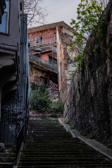 A wide outdoor staircase made of dark, weathered stone with multiple steps leading up to a building entrance in a narrow, urban street. The stairs are flanked by metal handrails on both sides, and the